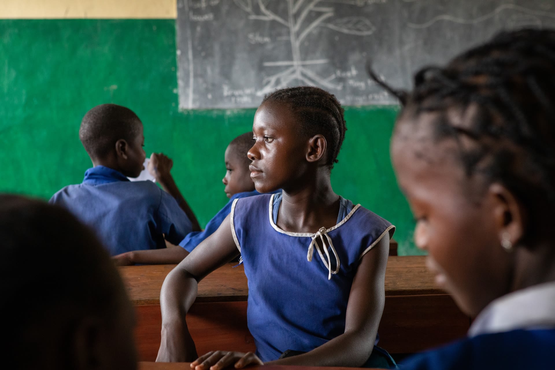 Tenneh, in a blue dress, sits in a classroom in front of a blackboard at her school in Pujehun district, Sierra Leone